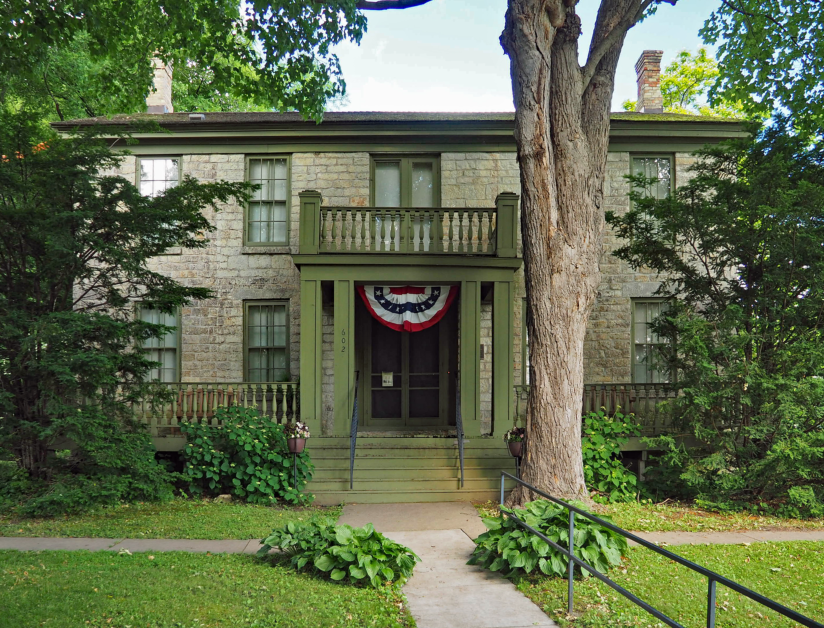 Warden’s House Museum stone exterior and green porch in Stillwater, Minnesota; a large tree partly obscures the facade.